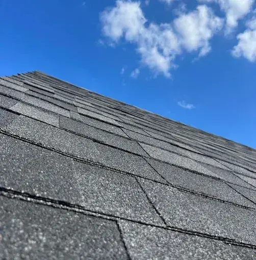 Dark asphalt shingle roof against a bright blue sky with fluffy white clouds.