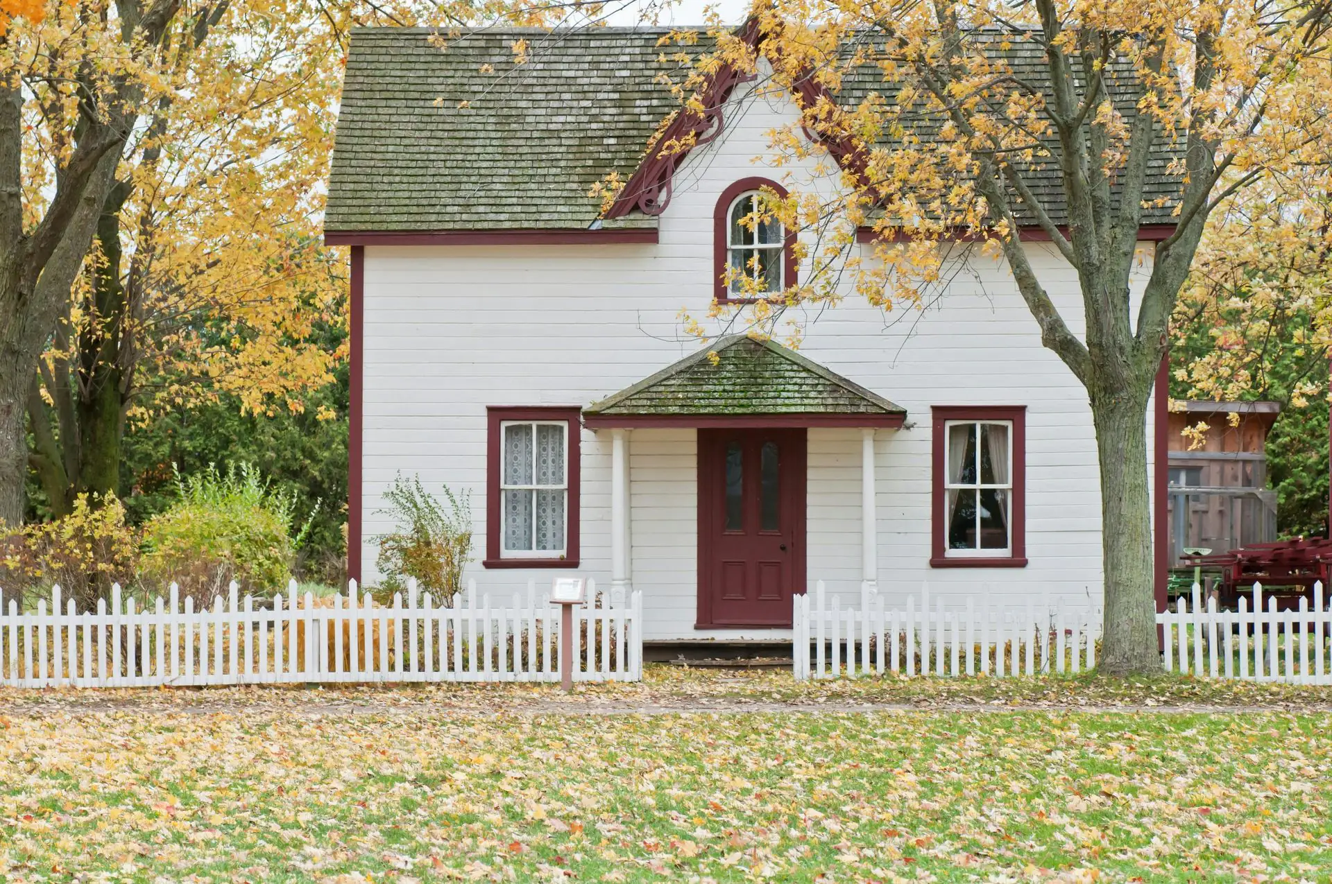 White farmhouse with a red door behind a white picket fence and autumn trees