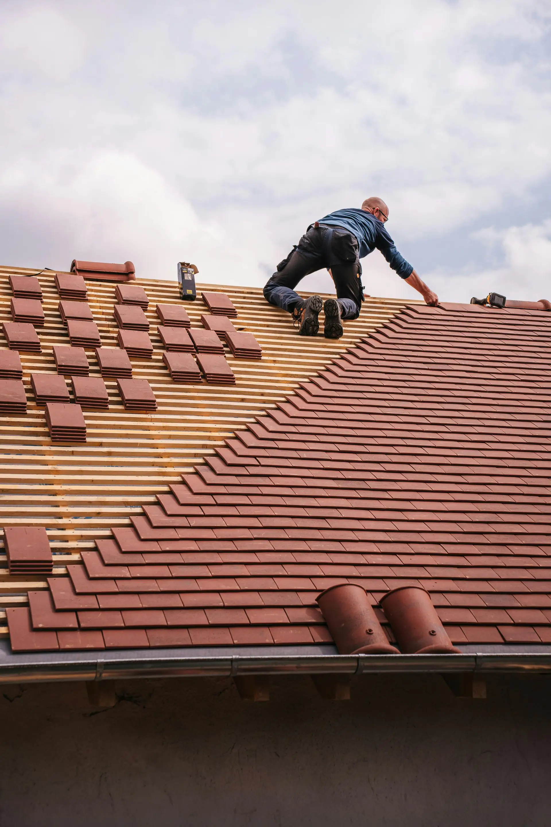 Roofer kneeling on a pitched roof, laying red clay tiles over exposed battens