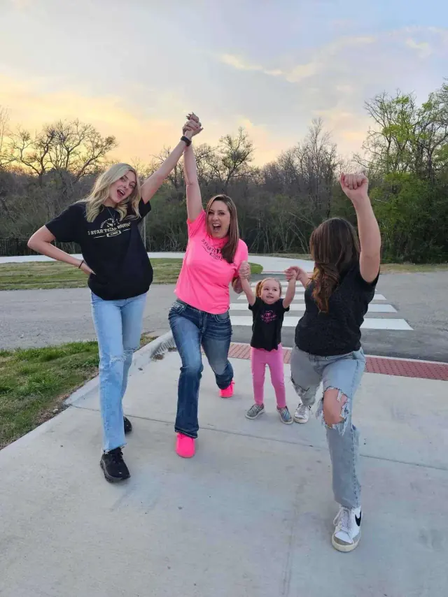 Four people cheering and raising arms on a sidewalk: a blonde teen, a woman in pink, a child, and a teen in torn jeans.