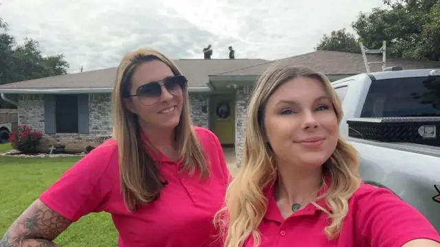 Two women in pink shirts smile in front of a house. Workers on the roof, truck nearby.