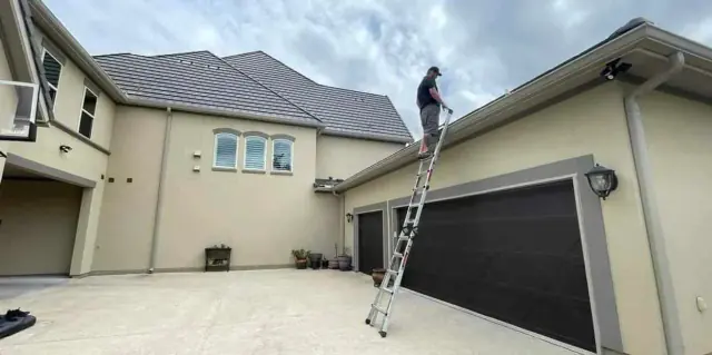 Person on a ladder cleaning a gutter on a light-colored house with a striped roof. Cloudy sky in the background.