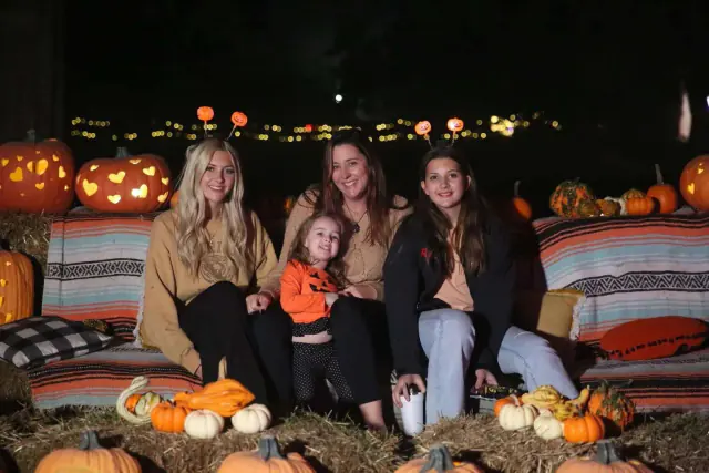 Four people seated on hay bales at night, surrounded by pumpkins and carved pumpkin lights.