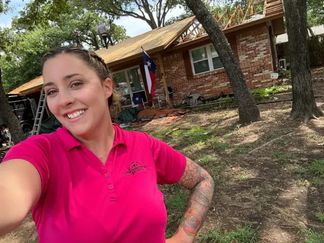Woman in pink shirt smiles, taking a selfie in front of a house with roofing work in progress.