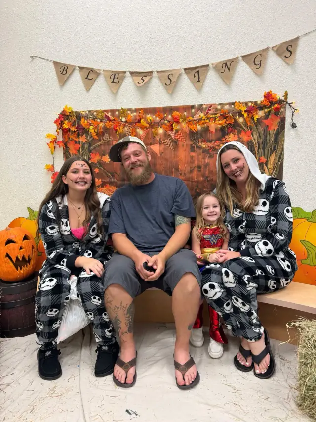 Family in skull-print pajamas poses on bench in front of autumn backdrop, pumpkins nearby.