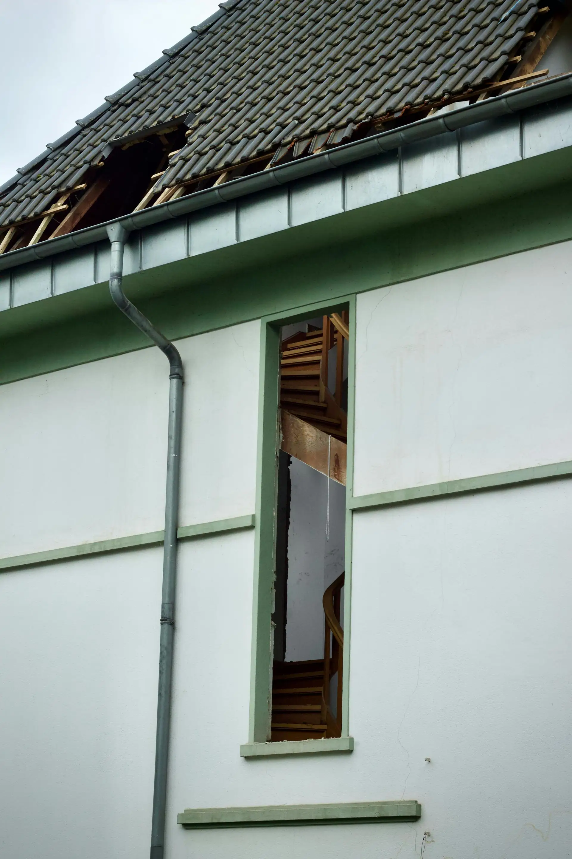 Storm-damaged roof with missing sections above a pale green wall and metal downspout