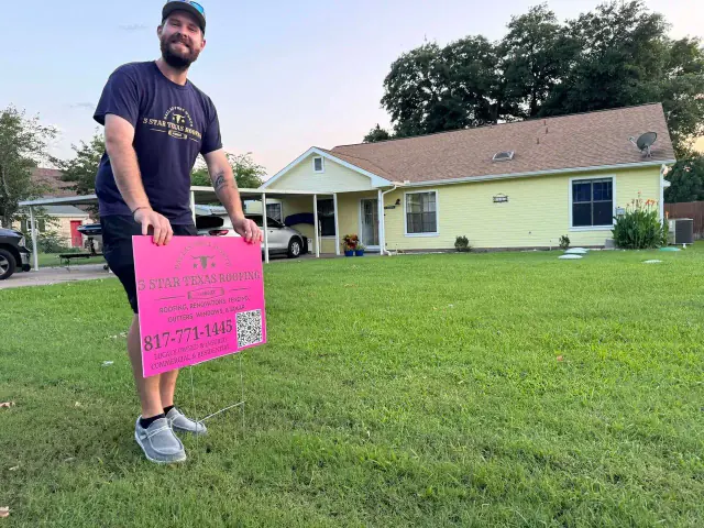 Man holding pink sign in front of yellow house; green lawn.